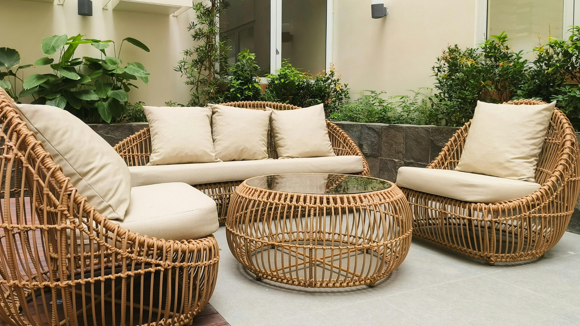 A modern outdoor patio with wicker furniture, including a sofa, two armchairs with beige cushions, and a round glass-top wicker table set atop well-kept pavers that highlight the importance of regular paver maintenance, surrounded by green plants and neutral walls.