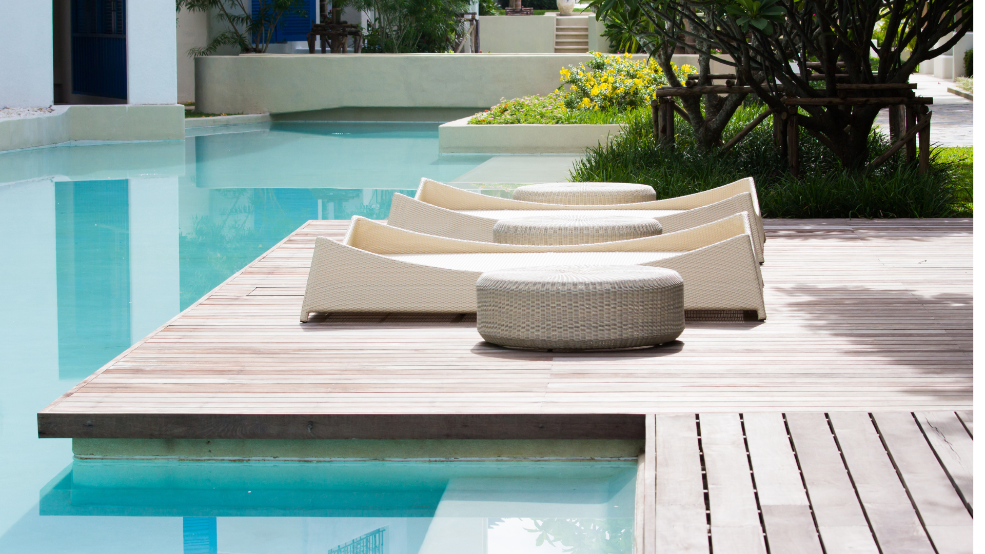 Three modern lounge chairs and two round ottomans sit on a wooden deck beside a serene, light blue swimming pool—an inviting oasis that highlights the difference between professional paver installation vs. DIY for your outdoor space.