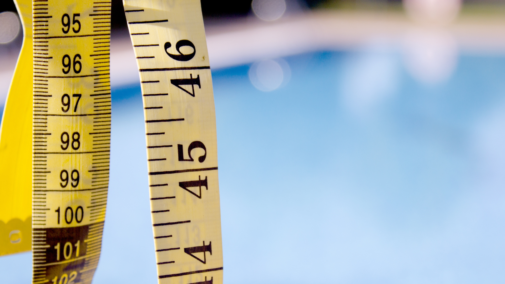 A close-up of a yellow measuring tape with black numbers, hanging in front of a blurry blue background, possibly a swimming pool—perfect for when you need to measure your pool for the perfect Loop-Loc cover fit. The tape shows measurements in inches.